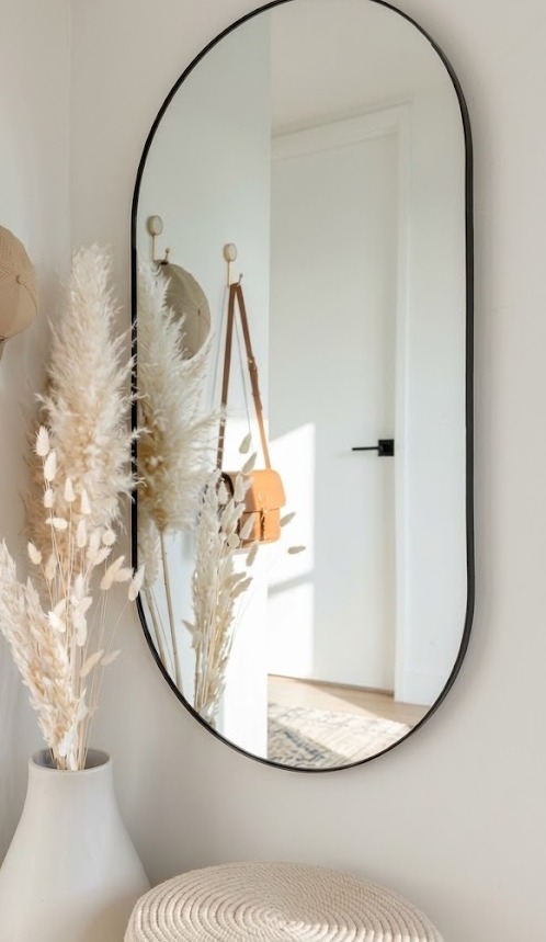 A tall oval mirror reflecting the entryway, beside a vase of dried pampas grass and a woven pouf